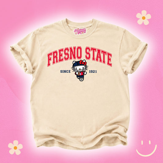 A beige short-sleeve t-shirt featuring a red 'FRESNO STATE' graphic and a Hello Kitty illustration on the front, displayed against a pink background with decorative flower and smiley face graphics.