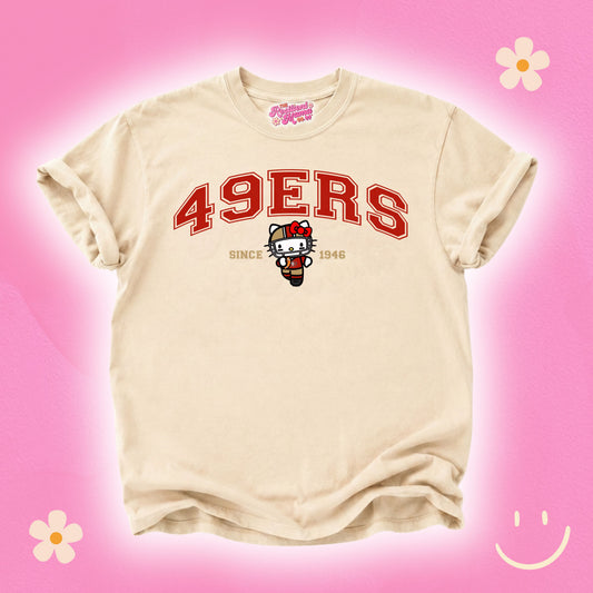 A beige t-shirt featuring a red '49ERS' graphic and a Hello Kitty character on the front, displayed against a pink background with decorative flower and smiley face graphics.