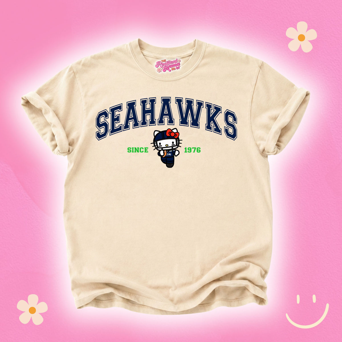 A cream-colored short-sleeve t-shirt featuring a navy blue 'SEAHAWKS' graphic with a cartoon cat mascot and 'SINCE 1976' printed on the front. The shirt is displayed against a bright pink background decorated with simple flower and smiley face illustrations.