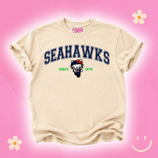 A cream-colored short-sleeve t-shirt featuring a navy blue 'SEAHAWKS' graphic with a cartoon cat mascot and 'SINCE 1976' printed on the front. The shirt is displayed against a bright pink background decorated with simple flower and smiley face illustrations.